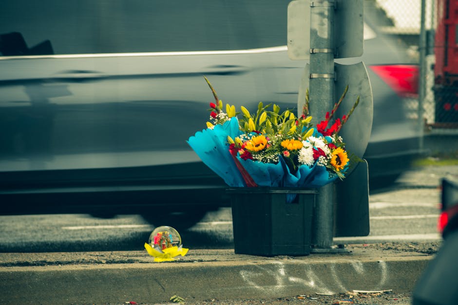 A vibrant floral arrangement of fresh flowers, including sunflowers, red and yellow blooms, and white daisies, wrapped in blue paper and placed in a black plastic container on the pavement beside a st