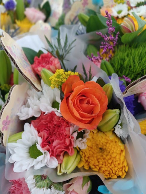 A vibrant floral bouquet featuring an orange rose as the focal point, surrounded by white daisies, pink carnations, yellow chrysanthemums, and purple lavender sprigs. The flowers are arranged with gre