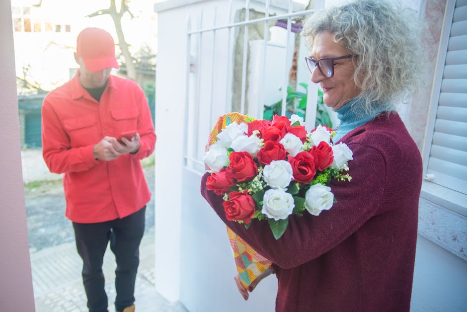 A woman with curly gray hair and glasses holds a bouquet of fresh red and white roses wrapped in brightly patterned paper, smiling as she receives the floral gift outside a house. In the background, a