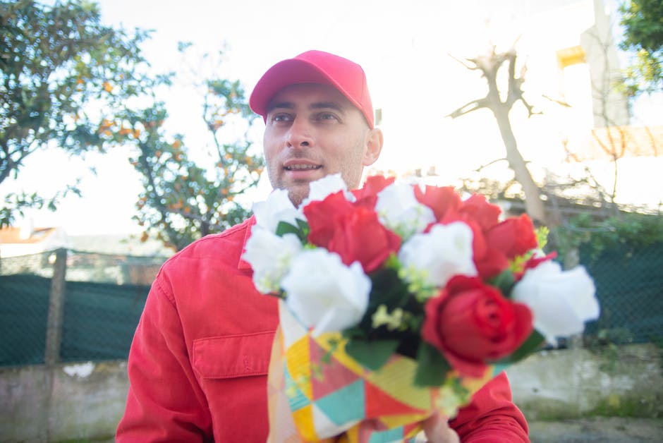 A male florist wearing a red cap and matching red jacket holds a colorful bouquet of fresh flowers, including red and white roses and other blossoms, wrapped in patterned paper. He is outdoors in a ga