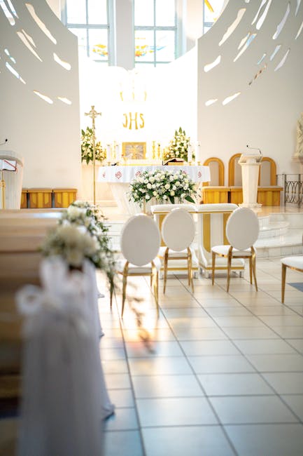 A bright, minimalist church interior prepared for a wedding ceremony, featuring an altar decorated with a large white floral arrangement with roses, peonies, and greenery, placed on a white cloth-cove