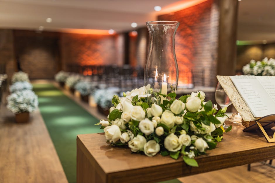 A floral arrangement of fresh, white roses with green foliage is placed on a wooden table, complemented by a tall, clear glass vase and an open, ornate book. The background features a softly lit indoo
