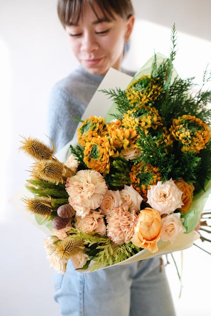 A young person with short hair and a light grey top holding a large bouquet of fresh flowers. The arrangement includes yellow, orange, and peach roses, yellow and green chrysanthemums, and dried fenne
