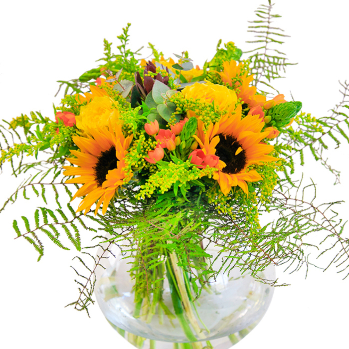 Bursting with seasonal vitality, this vibrant bouquet is a cheerful homage to Thamesmead's open skies and lush parks. Towering sunflowers - their faces framed in vivid yellow and offset by chocolate-brown centers - bring to mind sun-dappled afternoons by Southmere Lake. Intertwined with them, creamy yellow garden roses bloom with layered precision, each petal unfolding in a soft spiral of elegance and warmth. Sprays of bright red hypericum berries add playful dimension, their gleam standing out among the seas of verdant foliage. Delicate fern fronds flow down the sides of the arrangement, expertly mimicking wild flora found near the Thames, while sprigs of goldenrod sparkle with miniature yellow flowers. Slender green spikes and bluish eucalyptus leaves complete the tableau, lending depth and an aromatic freshness. All are gathered in a spherical glass vase, where the stems are showcased as clearly as the wild grasses around Thamesmead's lakes and meadows. Gentle light accentuates the bouquet's textures, casting subtle shadows reminiscent of leafy canopies. With a subtle, inviting fragrance and a design steeped in local character, this arrangement is ideal for sharing joy on special occasions or sending a spontaneous expression of positivity in Thamesmead.